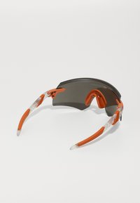 Ochelari de soare sportivi cu un lentilă mare gri, ramă din plastic portocalie și brațe transparente cu model. Include un pod nazal ajustabil.