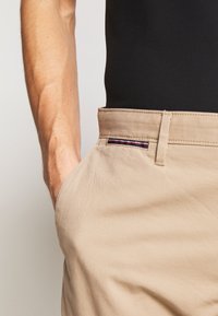 Pantalons en coton beige avec une texture lisse, dotés d'une poche latérale, de passants de ceinture et d'un accent décoratif rayé sur la taille.
