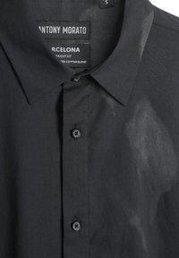 Zwarte shirt met korte mouwen van een katoenmix. Voorzien van een knopen sluiting aan de voorkant met een puntige kraag en logo labels zichtbaar binnenin de halslijn.