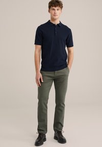 Camisa polo de manga corta en color azul marino con un patrón texturizado. Combinada con pantalones verde oliva y zapatos de cuero negros. El modelo está de pie frente a un fondo neutro.