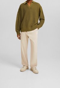 Personne portant un pull en maille vert olive, un pantalon ample beige et des baskets beiges, debout devant un fond uni gris clair.