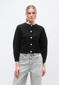 Μαύρο cropped ζακέτα κατασκευασμένη από απαλό πλεκτό υλικό, με μακριά μανίκια και στρογγυλή λαιμόκοψη, κλειστή με λευκά κουμπιά μπροστά.