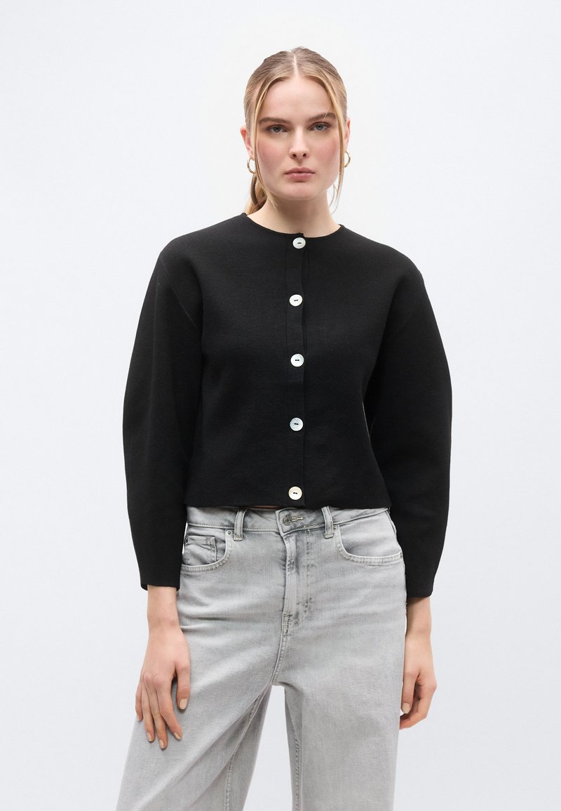 Μαύρο cropped ζακέτα κατασκευασμένη από απαλό πλεκτό υλικό, με μακριά μανίκια και στρογγυλή λαιμόκοψη, κλειστή με λευκά κουμπιά μπροστά.