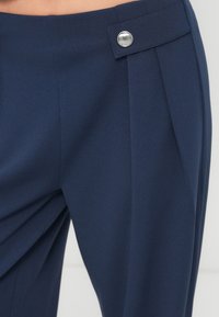 Σκούρα μπλε παντελόνια από λεία ύφασμα, με πτυχώσεις στο μπροστινό μέρος, πλαϊνή τσέπη και μεταλλικό κουμπί στη μέση.