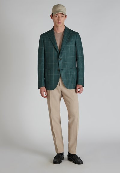 Zielona marynarka w kratę z brązowymi akcentami, beżowe spodnie i jasna czapka. Marynarka ma klapy z ząbkami i dwa guziki.