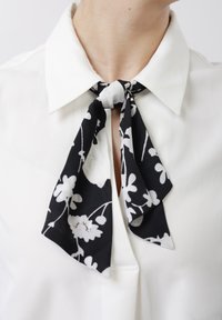 Λευκό πουκάμισο με γιακά και μαύρο-άσπρο φουλάρι με λουλουδάτο σχέδιο, δεμένο σε φιόγκο γύρω από τον λαιμό.