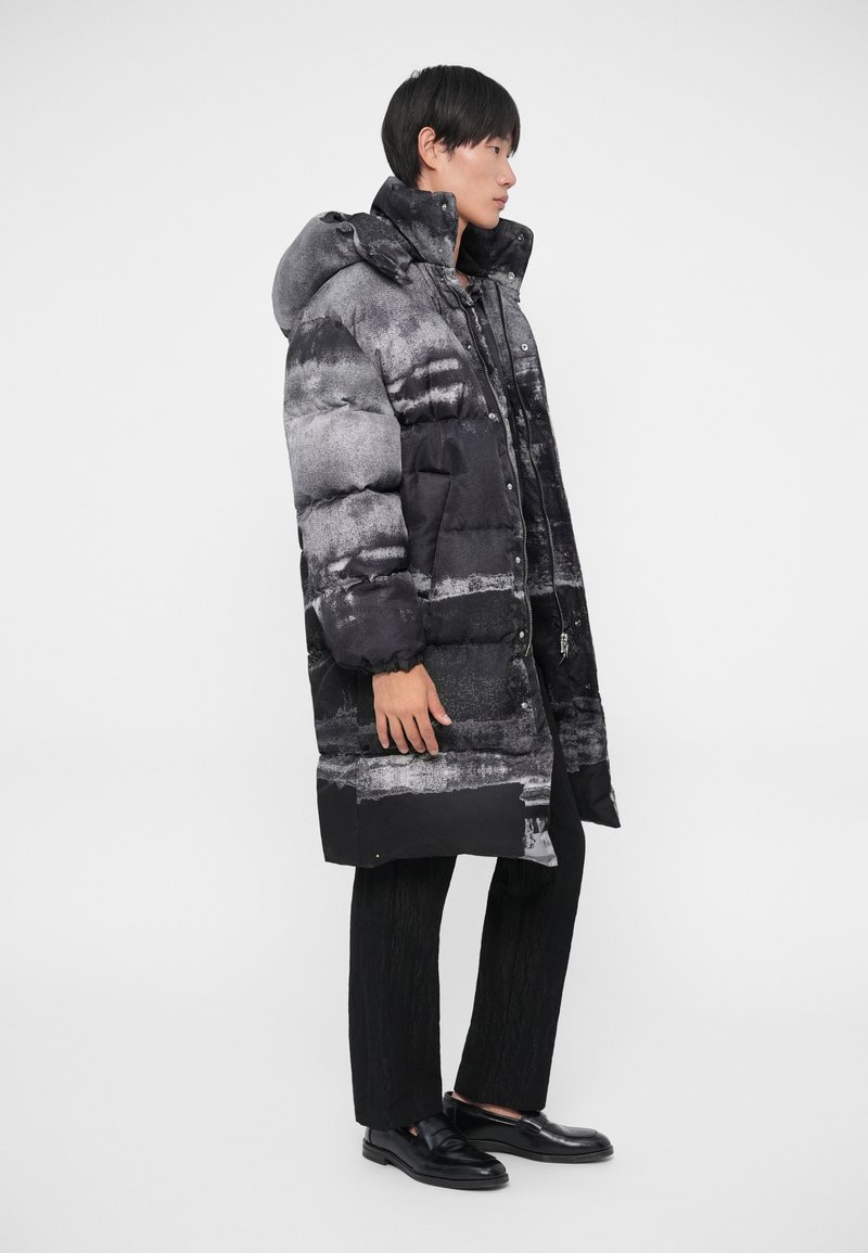 Sort og grå dunjakke med hætte, der har et stribet mønster og knaplukninger. Bæres over et sort outfit og elegante sko.