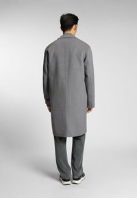Cappotto grigio di lana con taglio dritto, colletto a rever e dettagli sulle cuciture laterali, abbinato a pantaloni larghi scuri e calzature scure.