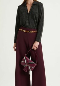 Donna che indossa un top nero a maniche lunghe, pantaloni bordeaux a gamba larga con cintura decorativa a catena, che tiene una borsa bordeaux con motivo e nappine.
