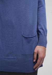 Pull en tricot bleu avec un col rond, des manches longues et une poche à l'avant. L'ourlet est côtelé et la texture semble douce.