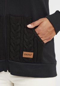 Čierna mikina na zips s pletenými vreckami, s hnedým značkovým znakem. Hranaté manžety a pás, vyrobené z mäkkého materiálu.