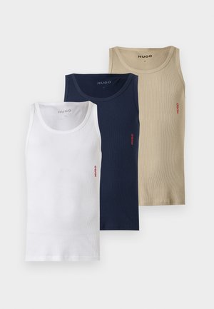 Erwachsener schwarzer Mann steht barfuß, trägt ein enganliegendes weißes ärmelloses Tanktop und dunkelblaue Boxershorts vor einem einfachen weißen Hintergrund.