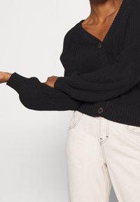 Cardigan nero lavorato a maglia con scollatura a V, maniche lunghe e chiusura con bottoni. Il tessuto presenta una texture a costine e polsini aderenti. Abbinato a pantaloni chiari.