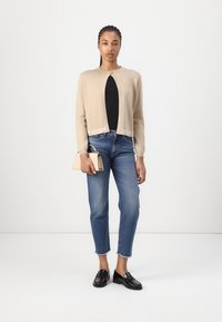 Beige neulottu lyhyt cardigan, jossa on pyöreä kaula-aukko ja logotyyppinen yksityiskohta, yhdistettynä tummansinihin farkkuihin ja mustiin loafer-kengiin, pitäen pientä beigeä käsilaukkua.
