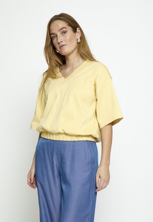 Kvinde med langt lyst brunt hår iført gul V-hals kortærmet top og løstsiddende blå bukser, stående mod en ensfarvet hvid baggrund.