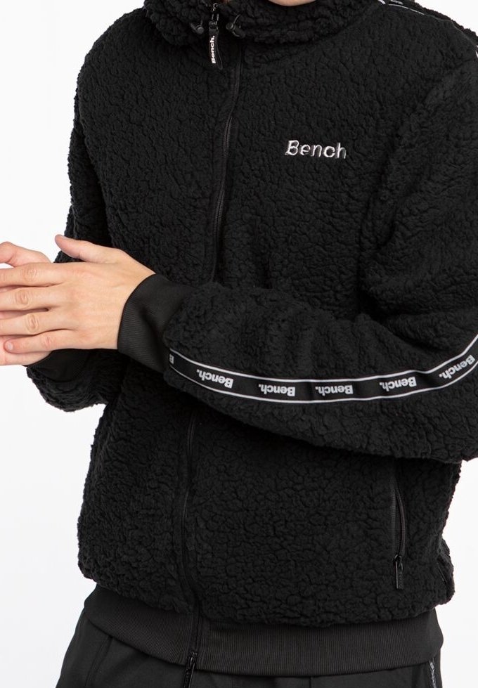Person trägt eine schwarze, strukturierte Jacke mit Reißverschluss und "Bench"-Logos auf Brust und Ärmeln, Hände vors Körper verschränkt.
