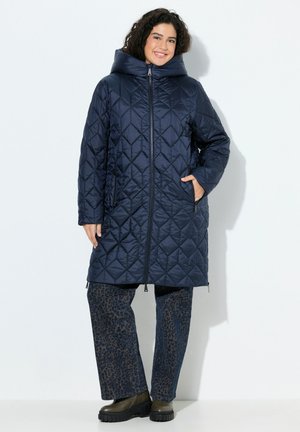 Abrigo largo acolchado azul marino con una gran capucha, cremallera frontal y dos bolsillos laterales; combinado con pantalones estampados y botas negras gruesas.