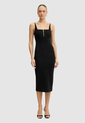 Mujer con un vestido midi negro ajustado con tirantes finos y cremallera frontal, combinado con tacones negros de punta abierta y pendientes de aro.