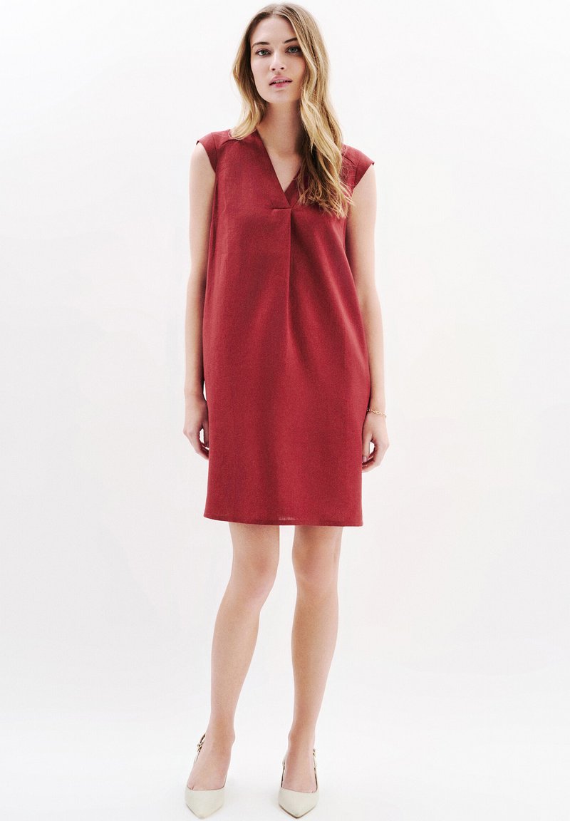 Punainen hihaton mekko, jossa on V-pääntie, rento istuvuus ja teksturoitu kangas. Mekossa on hienovaraisia laskoksia rinnassa ja se ulottuu polven ylle.