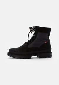 Bota negra de ante y textil con cordones, que presenta un cuello acolchado, suela de goma gruesa y texturas contrastantes en la parte superior. Su diseño elegante mejora la durabilidad.