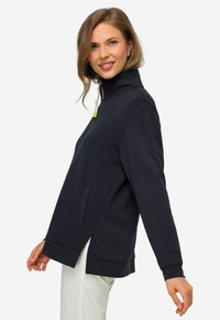 Sudadera azul marino con cremallera y cuello alto, aberturas laterales y bolsillos frontales. Fabricada en una tela suave con una textura lisa y un diseño minimalista.