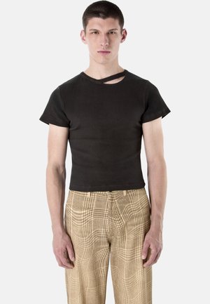 Junger Mann, der ein schwarzes kurzes Oberteil mit einem diagonalen Ausschnitt nahe dem Kragen und beige karierte Hosen trägt, steht aufrecht.