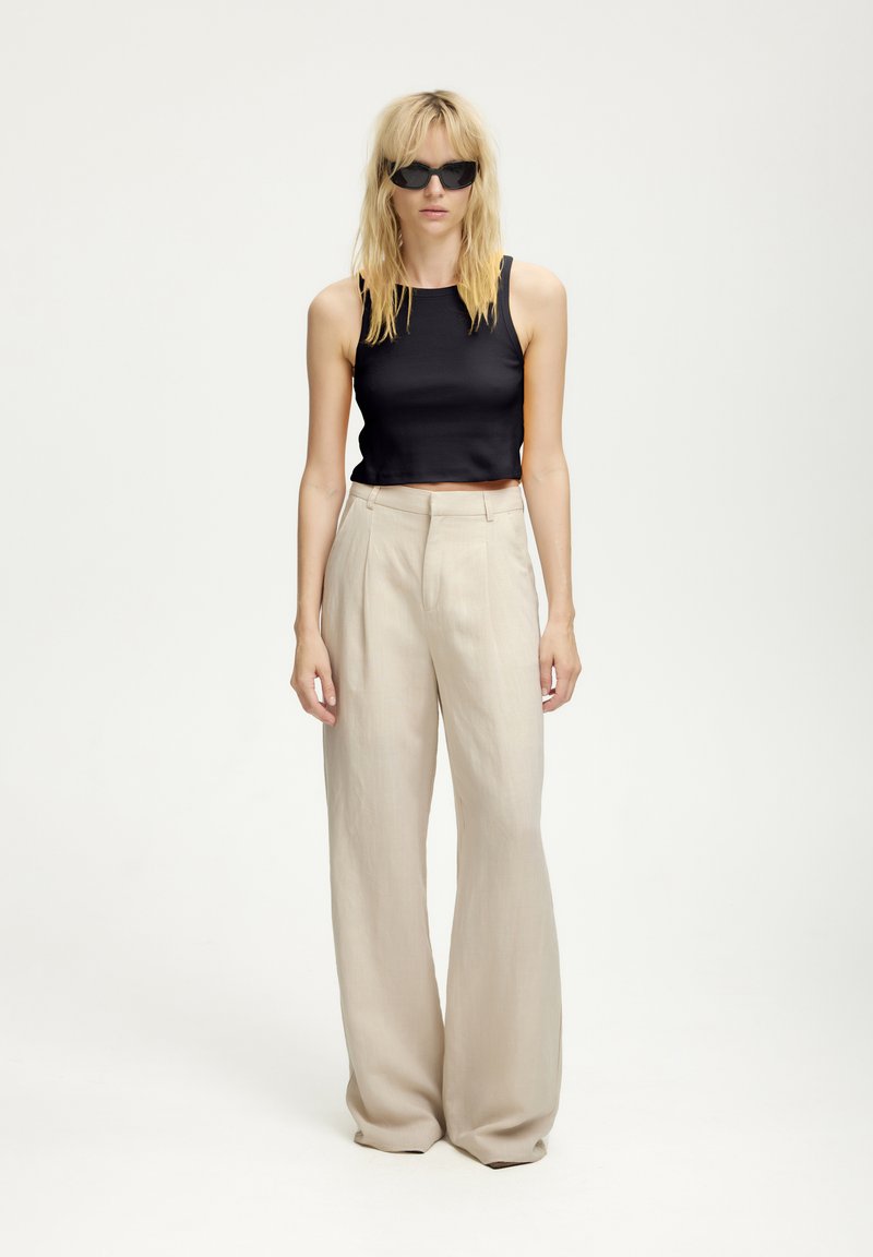 Čierny rebrovaný crop top v kombinácii s béžovými širokými nohavicami s vysokým pásom. Modelka má tiež na sebe čierne slnečné okuliare. Jednoduchý, moderný siluet.