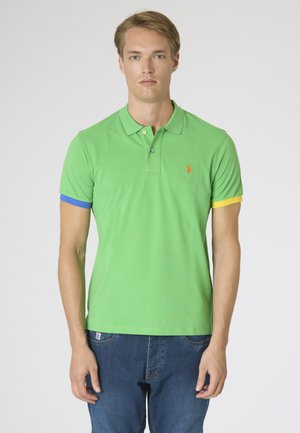 Giovane uomo che indossa una polo verde con polsini delle maniche blu e gialli e jeans blu, in piedi davanti a uno sfondo semplice.