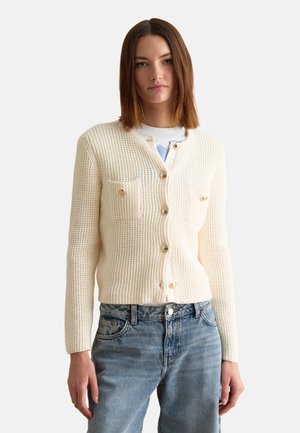 Donna che indossa un cardigan lavorato a maglia crema con bottoni e due tasche sul petto, abbinato a jeans di denim azzurro chiaro, in piedi su sfondo bianco.