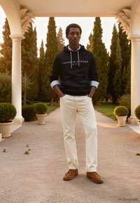 Sudadera con capucha negra con logo, camisa de botones azul claro debajo, pantalones crema y mocasines marrones. Ambientado en un jardín ajardinado.