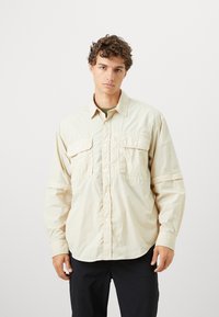 Camicia beige a maniche lunghe con due tasche anteriori, chiusura a bottoni e vestibilità leggermente più ampia. Tessuto leggero e texture liscia.