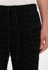 Črne trenirke z elastičnim pasom, ki imajo ponovljen vzorec besed "Versace" in "Jeans" v črni barvi na črni podlagi. Z vrvico za zapiranje.