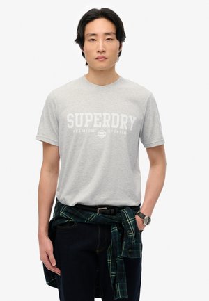 Szara koszulka z krótkim rękawem z białym logo "SUPERDRY", w zestawie z ciemnymi dżinsami i zieloną koszulą w kratę związaną w talii.