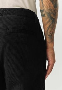 Black corduroy pants feature vertical ribbed texture, rear pocket, and a fitted waist. Slight sheen visible on the fabric.