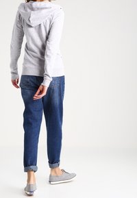 Sweat à capuche gris avec capuche, associé à un jean bleu ample et des baskets grises. Texture visible sur le sweat, revers des manches du jean retroussés, et design plat des chaussures.