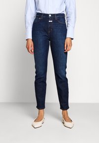 Pantalones vaqueros de mezclilla azul oscuro con cintura alta, ajuste entallado, diseño de cinco bolsillos y costuras contrastantes en color naranja, combinados con una camisa azul claro.