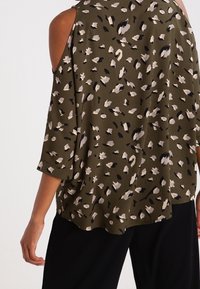 Femme portant un chemisier épaules dénudées vert olive avec des taches abstraites noires et beiges et un pantalon noir, vue de dos.