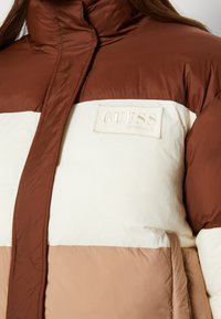 Kurtka puchowa w brązowym, kremowym i beżowym kolorze z blokowym wzorem. Posiada wysoki kołnierz oraz wytłoczoną naszywkę z logo "GUESS ORIGINALS".
