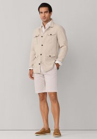 Homme debout portant une veste beige avec des poches à boutons, une chemise blanche, un short de couleur claire et des chaussures marron sans lacets, sur un fond neutre.