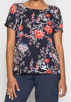 Femme portant un chemisier à manches courtes bleu marine à motifs floraux rouges, roses et bleu clair, assorti à un pantalon cargo bleu foncé.