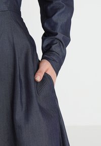 Robe bleu marine en tissu texturé, dotée de longues manches et d'une poche latérale. La coupe est cintrée à la taille et s'évase.