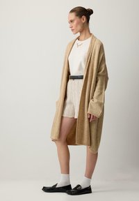 Beżowy sweter robiony na drutach, luźny krój, z długimi rękawami. Zestawiony z białą bluzką, wzorzystymi szortami, czarnymi mokasynami i białymi skarpetkami.