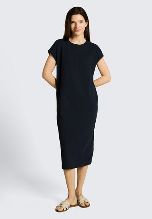 Kobieta w czarnej, krótkiej sukience do kolan i beżowych sandałach wsuwanych, stoi z rękami za plecami.