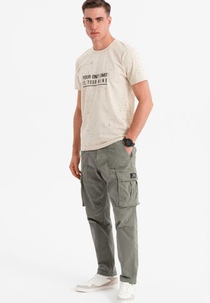 T-shirt beige con testo nero, abbinata a pantaloni cargo olive dal taglio loose fit con tasche laterali e un'etichetta, indossati con sneakers bianche.