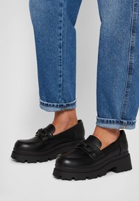 Mocasines de cuero negro con suela gruesa, que cuentan con una correa decorativa y un detalle texturizado. Combinados con jeans de mezclilla azul remangados.