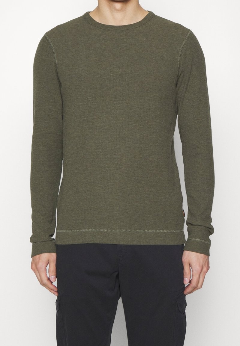 Homme portant un t-shirt à manches longues de couleur vert olive uni et un pantalon noir, debout devant un fond blanc uni.