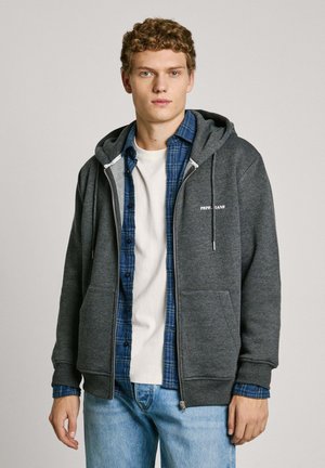 Grijze rits-hoodie met een kenguruzak, trekkoordkap, over een blauw geruit shirt en een wit T-shirt. Casual spijkerbroek.