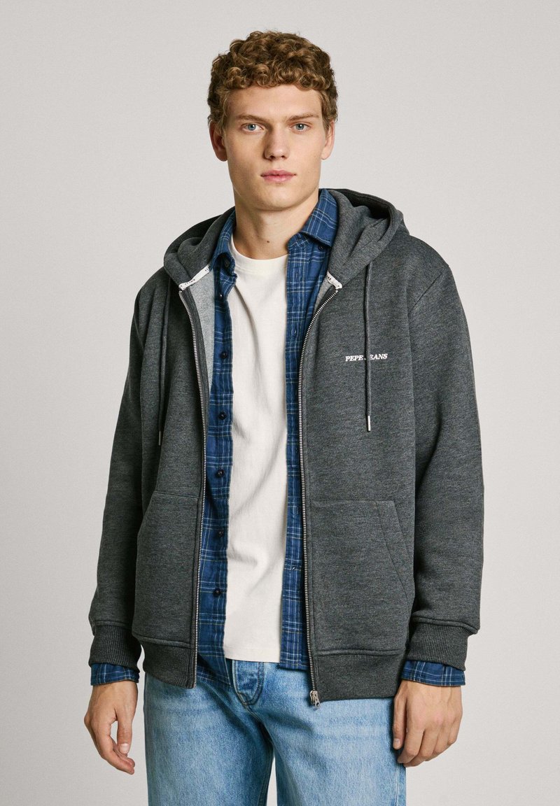Sudadera gris con cremallera y bolsillo tipo canguro, capucha con cordones, superpuesta a una camisa de cuadros azul y una camiseta blanca. Jeans de mezclilla casuales.