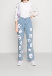Jeans di denim azzurro chiaro con motivi floreali ricamati bianchi. I jeans hanno una vestibilità rilassata e sono abbinati a un cardigan color crema.