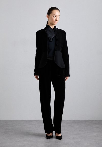 Jachetă neagră texturată cu detalii cu nasturi, asortată cu un top negru elegant și pantaloni din catifea negri. Modelul poartă tocuri negre.